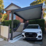 White car under a DIY cantilever carport near a brick house.