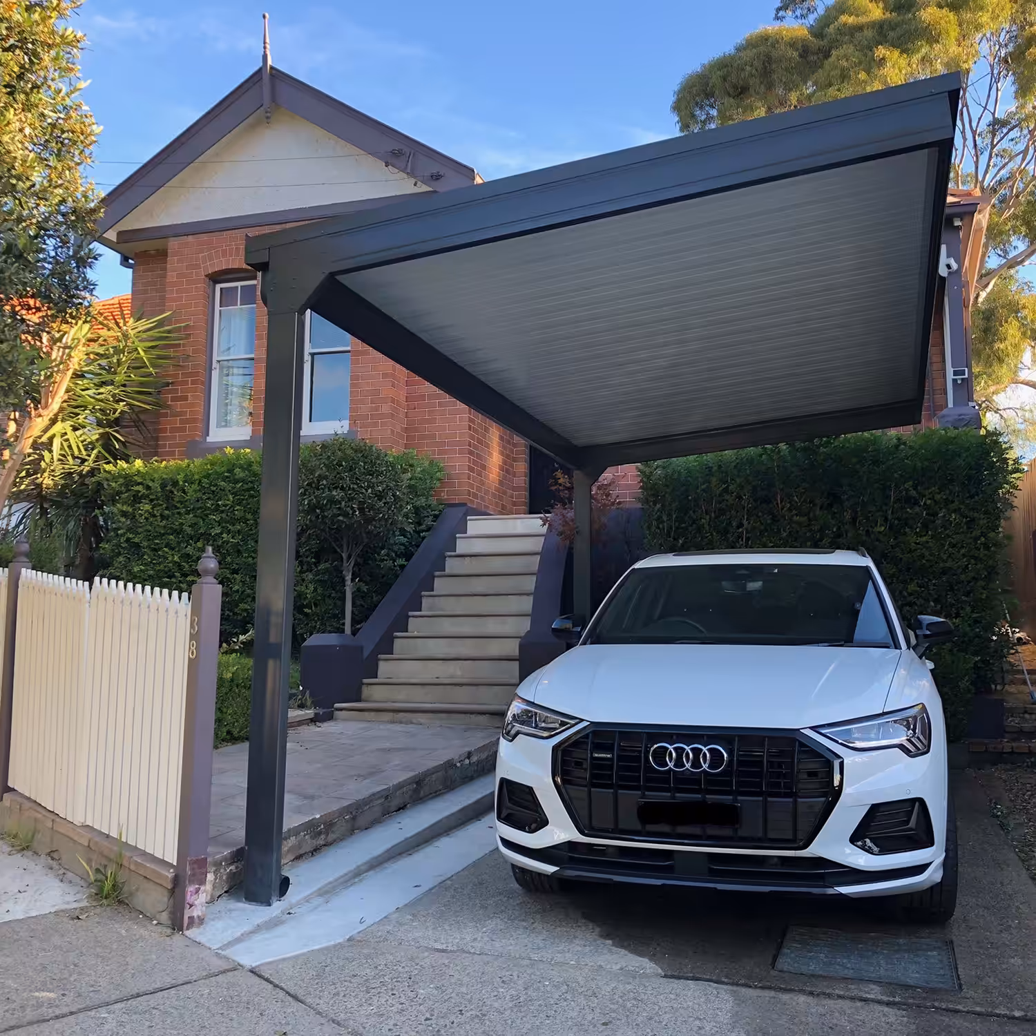 White car under a DIY cantilever carport near a brick house.
