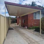 DIY freestanding flat roof carport beside brick house and garden.
